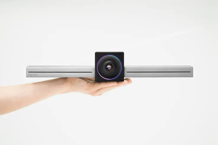 Webcam rests on an outstretched hand, featuring a black central camera with a glowing purple-blue ring and silver soundbar.