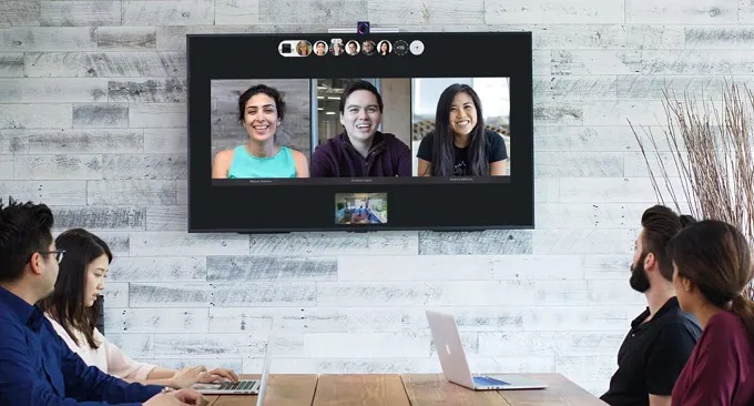 Colleagues gather around a table watching three participants on a wall-mounted video conference screen.