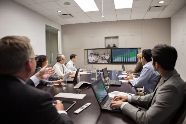 Business team seated around a conference table, viewing a wall-mounted presentation with bar charts and photos.