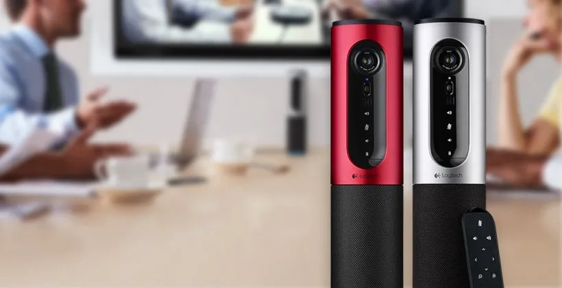 Video conferencing devices in red and silver stand beside a remote, with blurred people meeting around a table behind them.