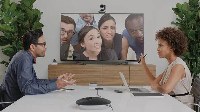 Colleagues discuss a video call at a conference table, with participants displayed on a wall-mounted screen.