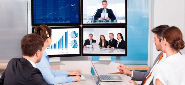 Employees view four computer monitors displaying charts and video conference screens around a desk.