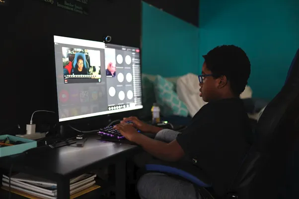 Person wearing glasses edits a video on a computer, seated at a desk with a keyboard, monitor, and stacked books.