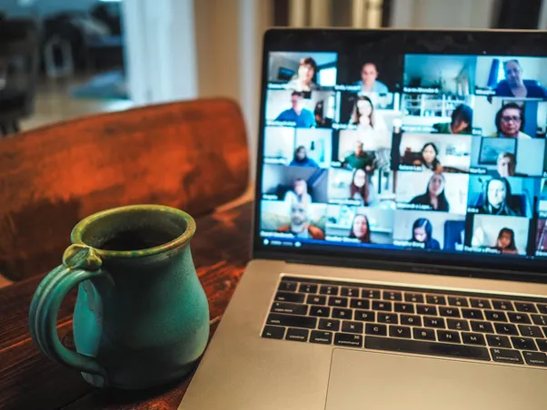 Laptop displays a grid of video conference participants beside a green mug on a wooden table.