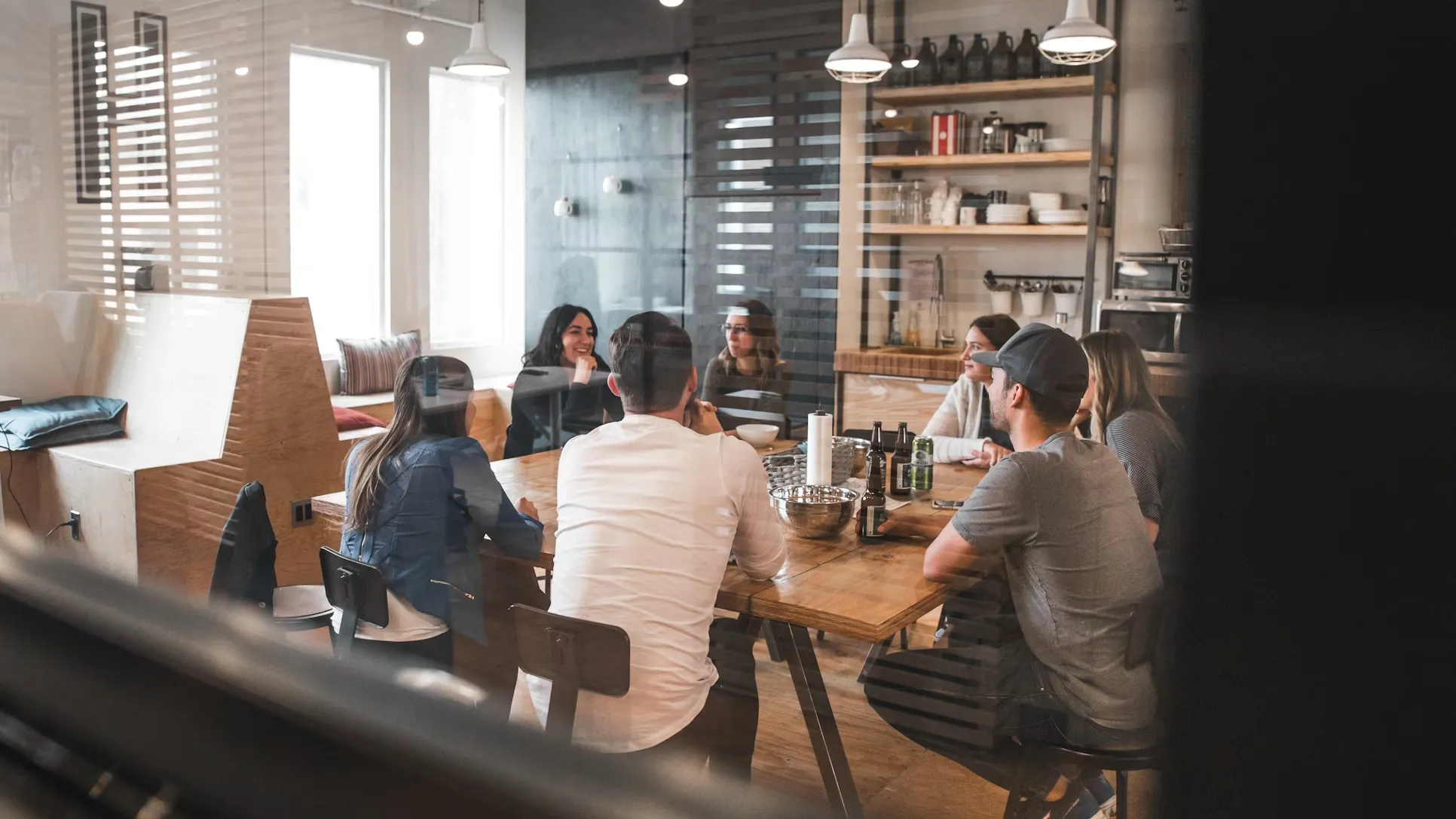 Diverse group of professionals having an informal meeting around a wooden table in a modern office kitchen area with open shelving and pendant lighting.