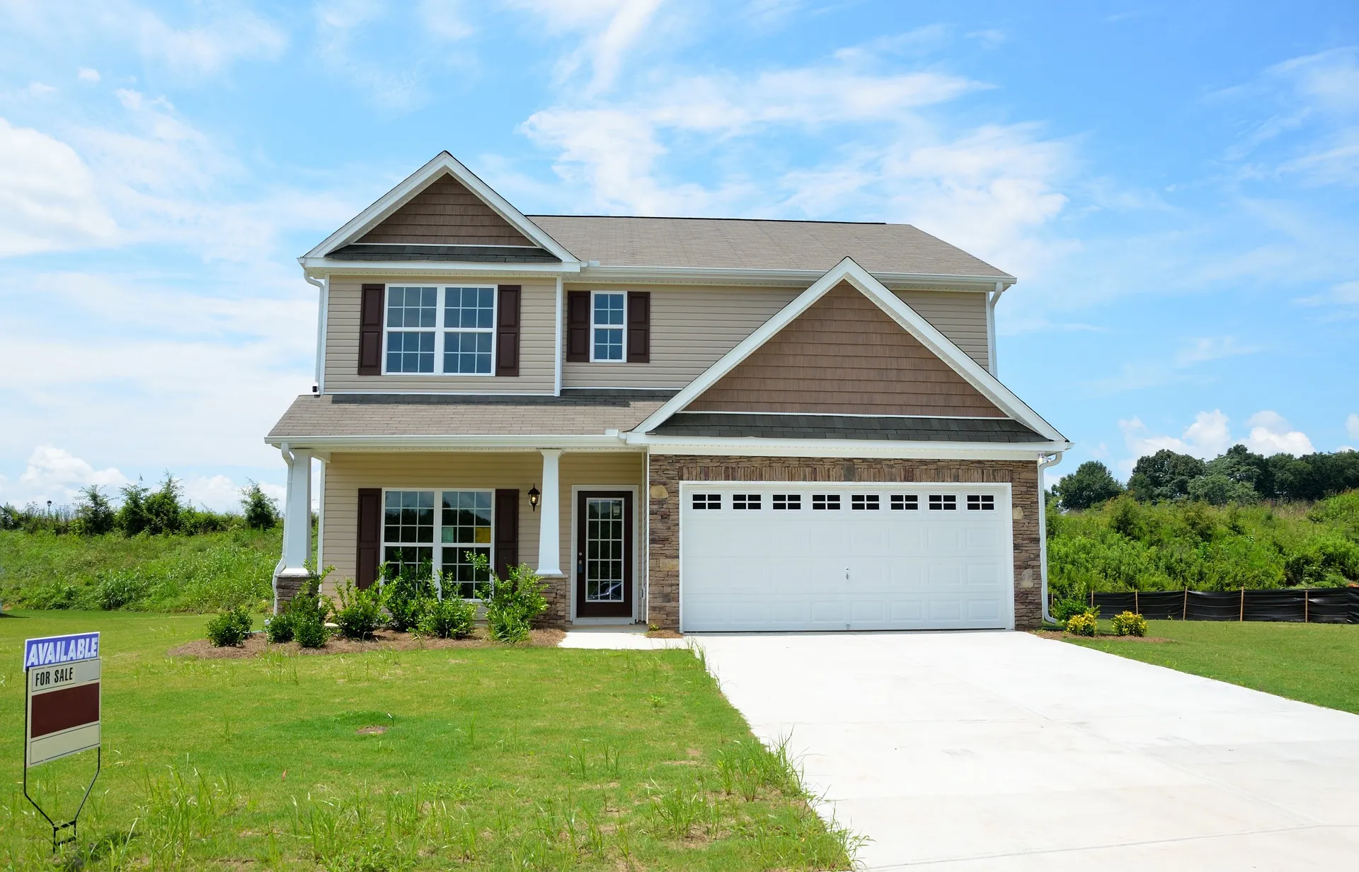 Two-story suburban house with beige siding, brown shutters, attached garage, and 'For Sale' sign on manicured lawn against blue sky