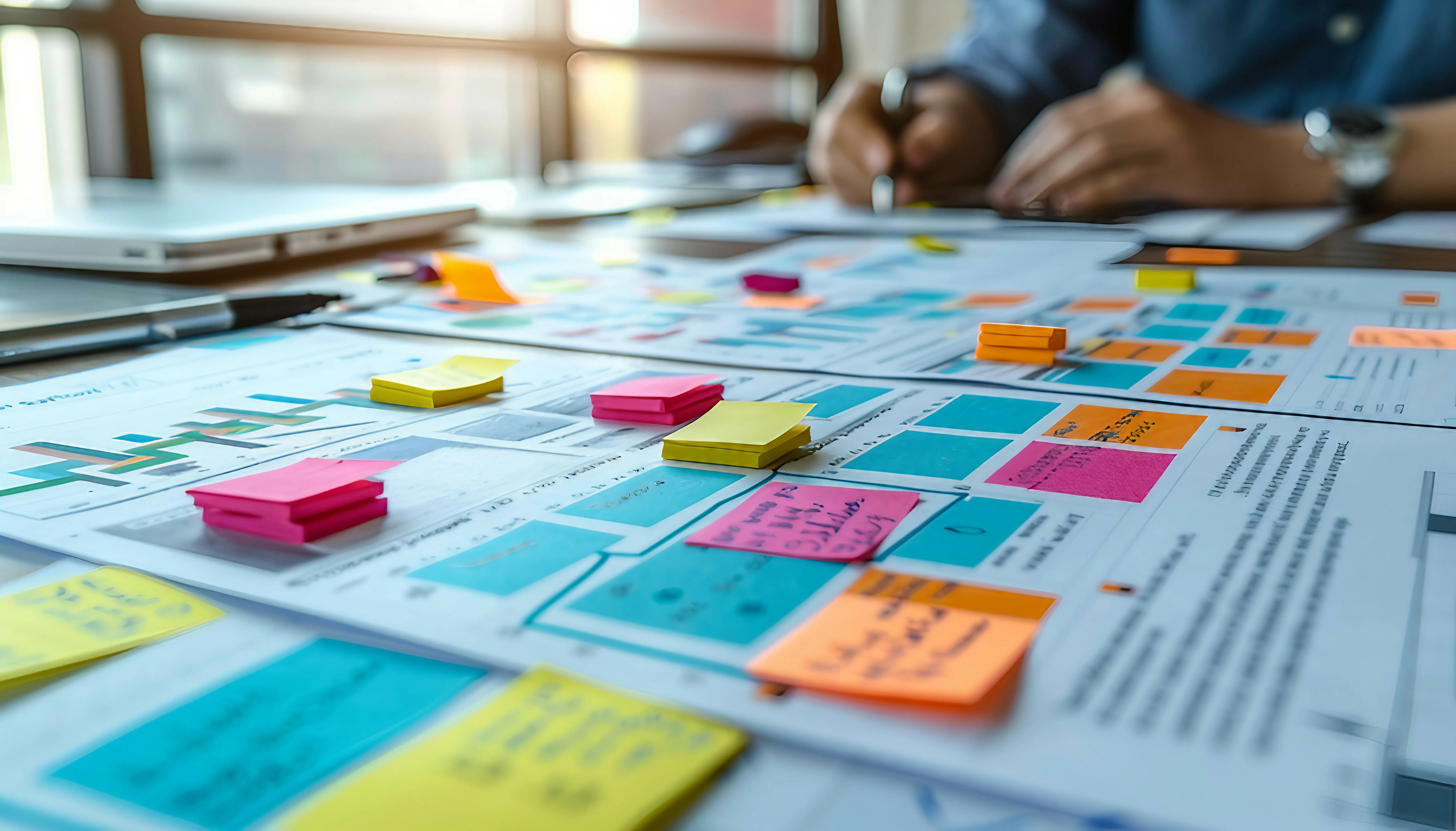 Desk with project planning documents covered in colorful sticky notes and a person writing in the background.