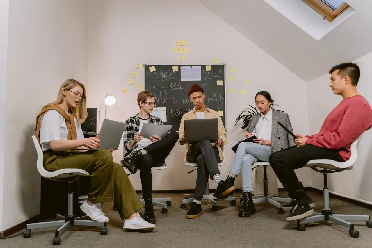 Five-person team collaborating in office with laptops and whiteboard.