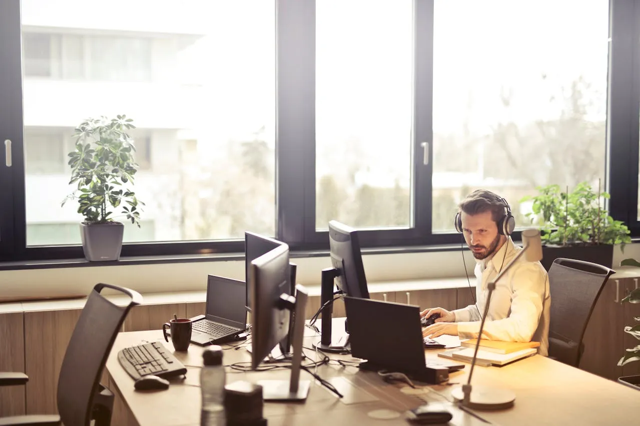 Man wearing headphones works on laptop and phone with plant.