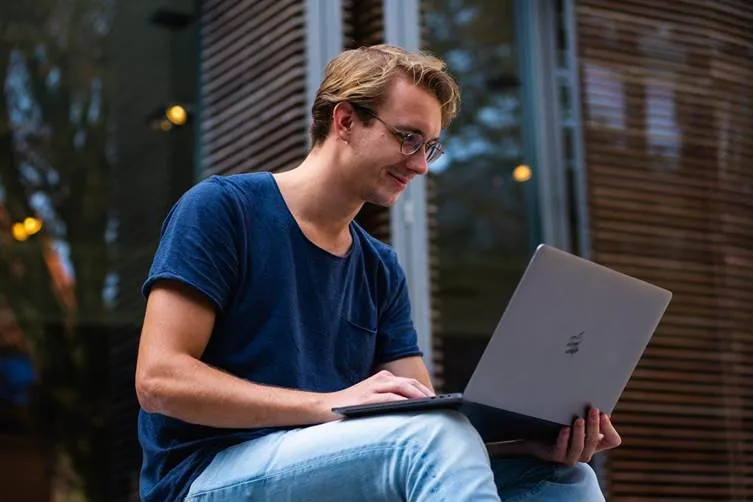 Young man using silver laptop outdoors near modern glass building.