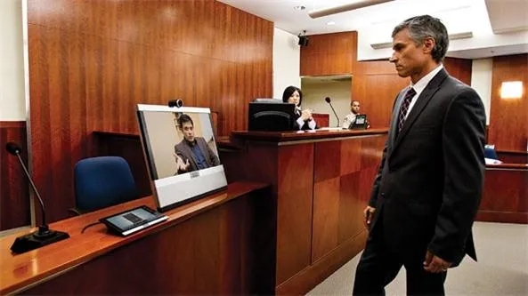 Courtroom features a suited man walking past a video monitor, microphones, wood-paneled desks, and seated officials.