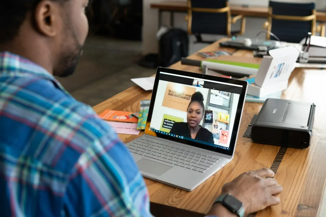 Man watches a video call on a laptop at a cluttered office table, with documents, chairs, and a printer nearby.