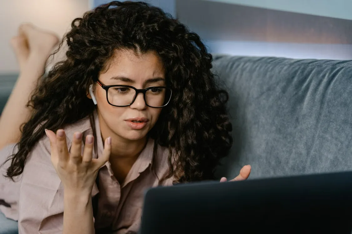 Woman wearing glasses gestures while looking at a laptop on a gray couch.