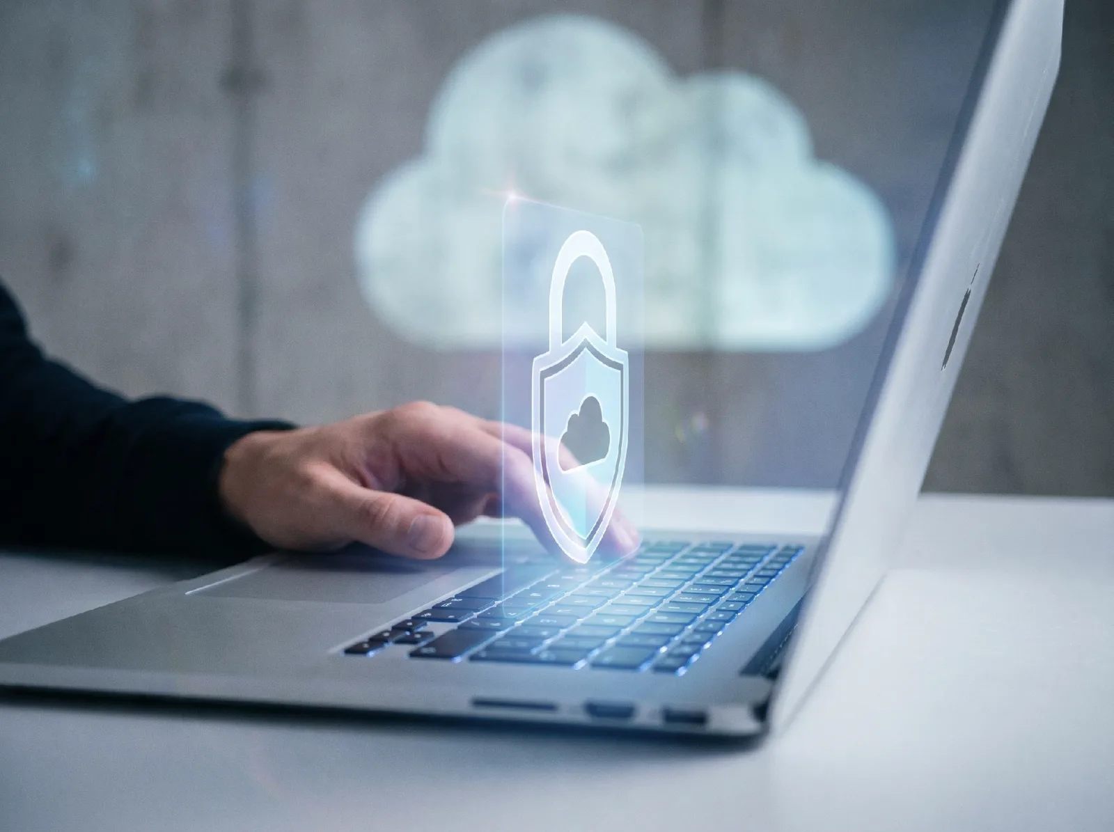 Laptop displays a glowing shield with a cloud symbol while a hand uses the keyboard, suggesting secure cloud computing.