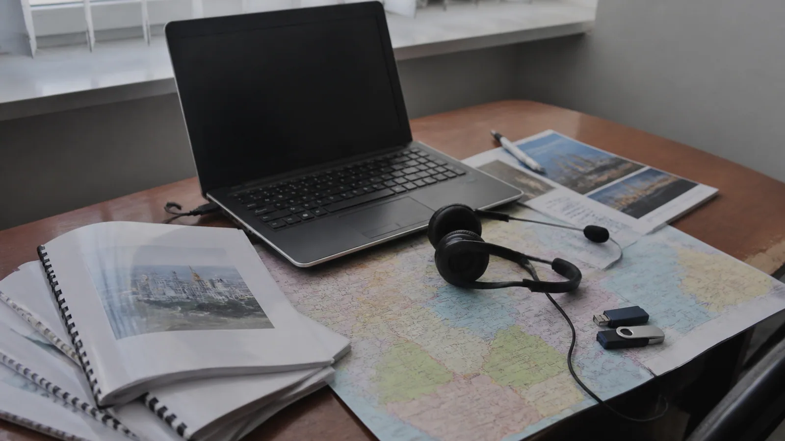 Laptop, headset, Cyrillic manuals, USB drives, reports, and a folded Eastern European map on a worn desk.