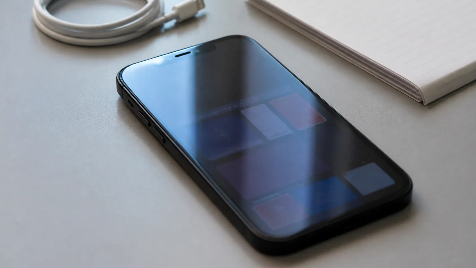 iPhone, charging cable, and notepad sit on a desk beneath cool light.