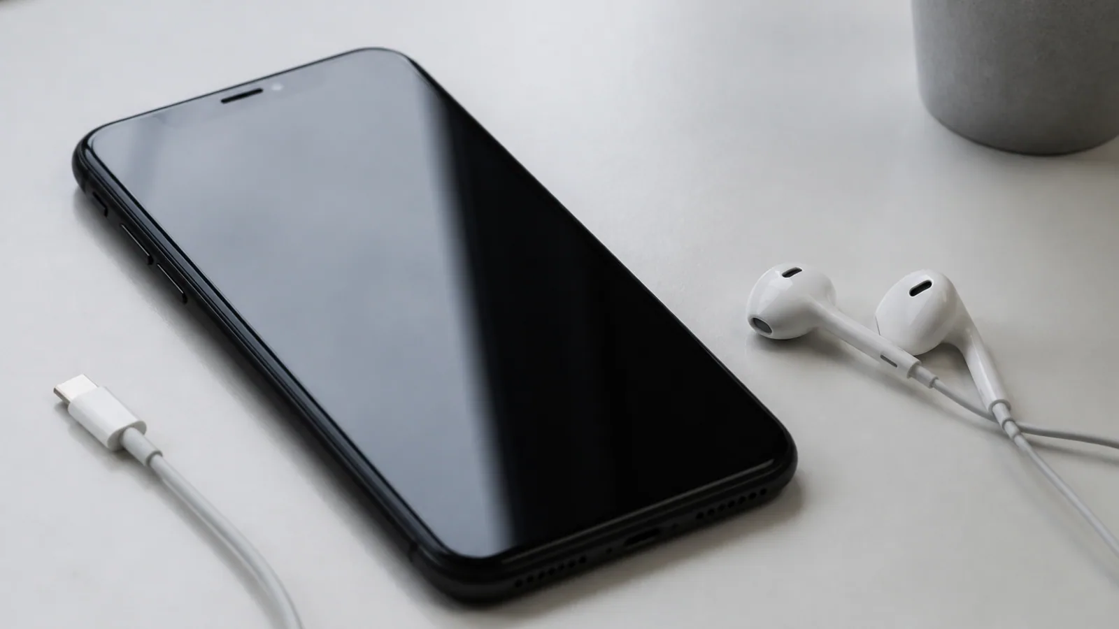 iPhone, charging cable, and earbuds rest on a pale desk beside a softly reflected screen.
