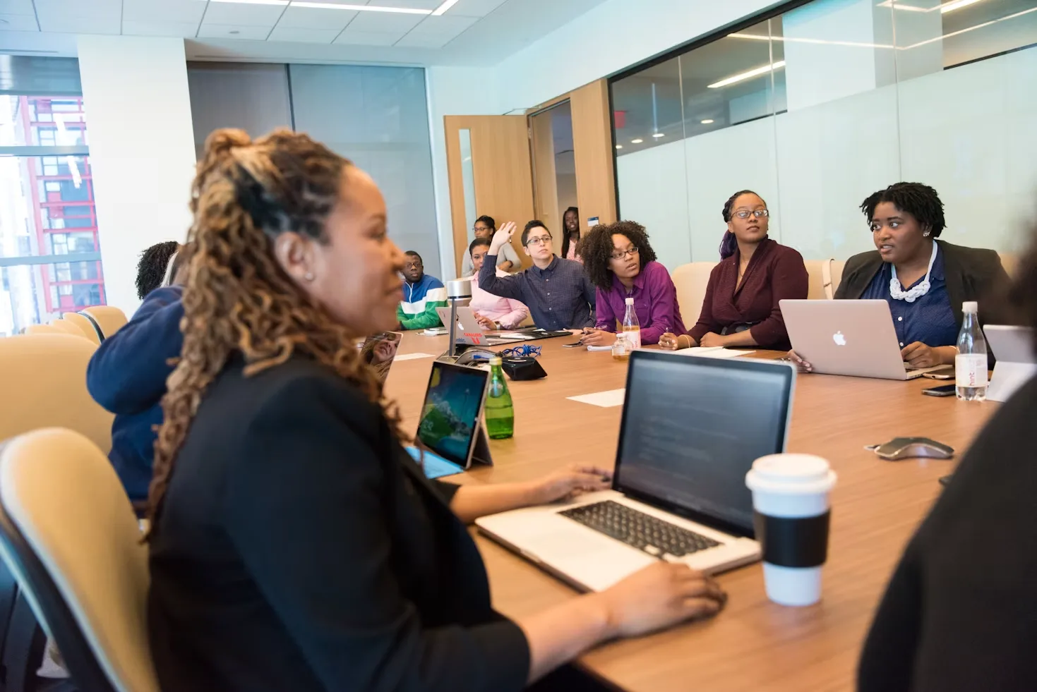 Colleagues gather around a conference table with laptops, listening and discussing in a bright glass-walled office.